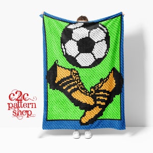 Pode incluir: Uma manta de crochê verde, azul e branca com uma bola de futebol e um par de chuteiras de futebol. A manta está sendo segurada por uma pessoa.