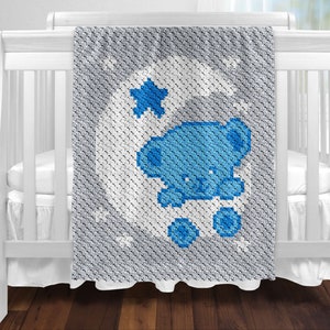 May include: A blue and white crocheted baby blanket featuring a blue bear on a crescent moon with a star. The blanket is draped over a white cot. The blanket has a grey border.