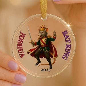May include: A clear circular acrylic ornament featuring a printed illustration of a rat wearing a gold crown, green military-style jacket, and red cape while holding a sword. The text JOSHUA, RAT KING, and 2025 are printed on the ornament.