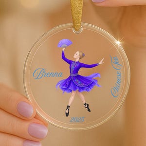 May include: Clear acrylic ornament featuring a ballerina in a purple dress and holding a fan. The ornament has the name "Brenna" and the text "Chinese Tea" in blue script. The year "2025" is also displayed. A gold ribbon is attached for hanging.