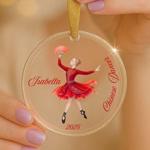 May include: Clear, round ornament with a gold ribbon. The ornament features a dancer in a red dress holding a fan. Text includes "Isabella", "Chinese Dancer", and "2025".