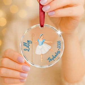 May include: A clear circular acrylic ornament hanging from a red ribbon. The center features a printed illustration of a shepherdess in a blue bodice and white skirt holding a staff. The text Lilly and Shepherdess 2026 is printed in blue.