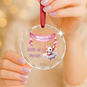 May include: A clear circular acrylic ornament featuring a pink banner with the text The Nutcracker, the text Addie as a Mouse, a small illustration of a mouse in a pink tutu, and the year 2026, suspended by a dark red satin ribbon.