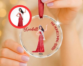 May include: A clear circular acrylic ornament hanging from a red ribbon. It features a printed illustration of a person in a red gown and white sash, with the text Kendall, FHS HOMECOMING QUEEN, and 2026 printed in red lettering.