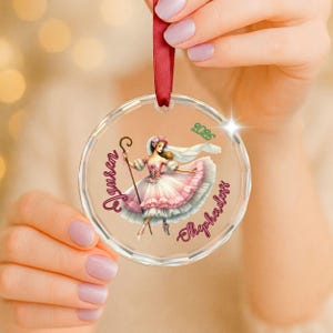 May include: A circular clear acrylic ornament featuring a printed illustration of a shepherdess in a ruffled pink dress holding a staff. The text "Lauren" and "Shepherdess" is printed in pink script, with the green year "2026" above the figure.