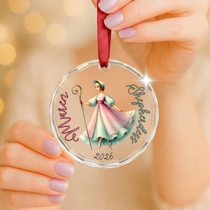 May include: A clear circular acrylic ornament featuring a printed illustration of a shepherdess in a flowing pastel dress holding a staff. The text "Maren" and "Shepherdess" is printed on the sides with the year "2026" at the bottom, suspended by a red ribbon.