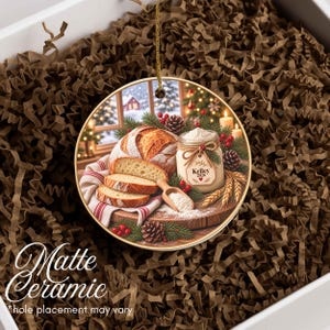 May include: A round ceramic ornament featuring a printed image of a loaf of sliced bread, a jar labeled Kelley 2026, pine cones, and evergreen branches. The ornament has a gold-colored string for hanging and a smooth, matte finish surface.