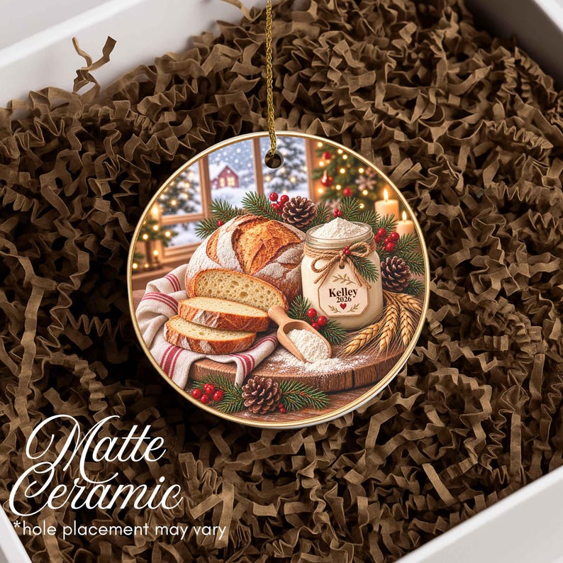 May include: A round ceramic ornament featuring a printed image of a loaf of sliced bread, a jar labeled Kelley 2026, pine cones, and evergreen branches. The ornament has a gold-colored string for hanging and a smooth, matte finish surface.