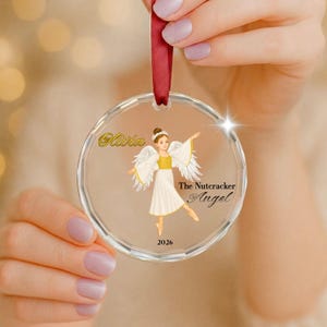 Peut inclure: Une décoration en verre transparent et circulaire ornée d'une illustration d'ange en robe blanche et dorée avec des ailes. Le texte indique Olivia, The Nutcracker Angel et 2026. Un ruban rouge est fixé au sommet pour la suspension.