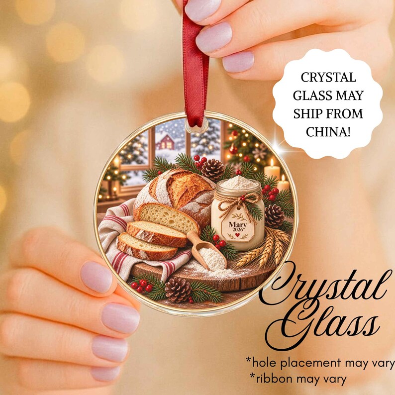 May include: A round glass Christmas ornament featuring a printed scene of a loaf of bread, sliced bread, a small jar labeled Mary 2026, a wooden scoop with flour, pine branches, and pinecones. The ornament hangs from a red satin ribbon.