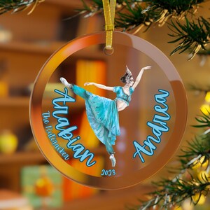 May include: A round glass ornament with a ballerina in a blue and gold costume. The ornament has the text "Arabian The Nutcracker" and "Andrea 2023" on it.