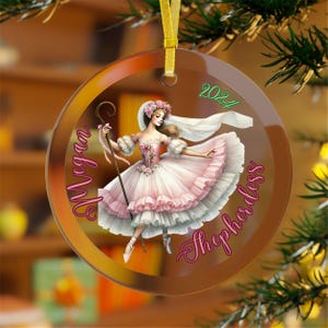 May include: A clear glass Christmas ornament with a pink and white ballerina figure holding a shepherd's crook. The ornament has the text "The Megan Shepherdess 2024" around the ballerina.