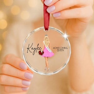 Peut inclure: Une décoration ronde en acrylique transparent suspendue par un ruban rouge. Le motif représente une silhouette blonde en robe rose tenant un bouquet rouge, avec le nom Kaylee, le texte Homecoming Queen et une écharpe WHS 2026 imprimés.