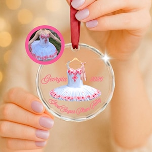 May include: A clear circular acrylic ornament hanging from a red ribbon. The ornament features a printed graphic of a white ballet tutu with pink floral accents, a pair of pink ballet slippers, the name Georgia, the year 2026, and the text Good Sugar Plum Fairy.