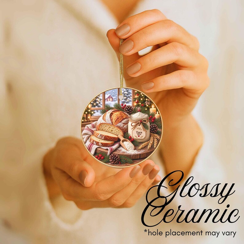 May include: A round glossy ceramic ornament featuring a festive illustration of a loaf of sliced sourdough bread, a small jar labeled Tara 2024, a wooden scoop with flour, pine branches, and red berries, all suspended by a thin gold string.