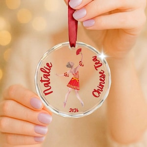 May include: A clear circular acrylic ornament featuring a printed illustration of a dancer in a red dress holding a fan. The text Natalie is printed on the left, Chinese Tea on the right, and 2026 at the bottom in red. A red ribbon is attached.