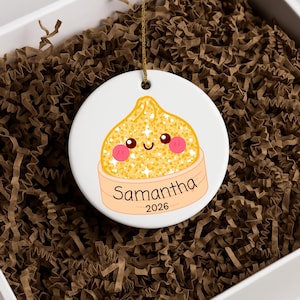May include: A round white ceramic ornament featuring a yellow dumpling illustration with a smiling face, pink cheeks, and sparkles. The dumpling sits in a light orange steamer basket with the name Samantha and the year 2026 printed in black text.