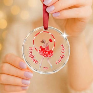 May include: A clear circular acrylic ornament featuring a red and white candy cane hoop graphic surrounding a ballerina in a red tutu. The text reads Meghan, Candy Cane, and 2026 in red script. A red ribbon is attached to the top for hanging.