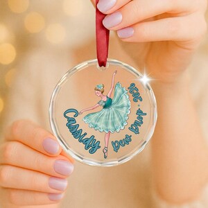 May include: A circular clear acrylic ornament featuring a printed illustration of a ballerina in a teal tutu. The text "Cassidy" and "Dew Drop 2026" is printed in blue script. A red satin ribbon is attached to the top for hanging purposes.