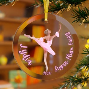 May include: A clear glass Christmas ornament with a pink ballerina in a pink tutu. The ornament has the text "Lydia" and "Sugar Plum Fairy" on it, along with the year "2024".
