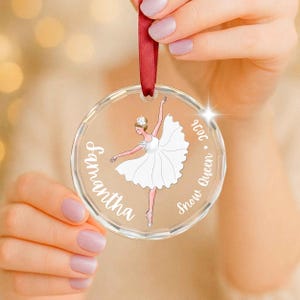 May include: A clear circular acrylic ornament featuring a printed illustration of a ballerina in a white tutu. The text Samantha and Snow Queen 2024 is printed in white script. A red satin ribbon is attached to the top for hanging purposes.