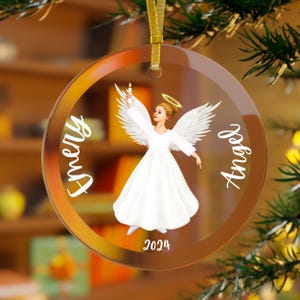 May include: A round, clear glass Christmas ornament with a gold ribbon. The ornament features a white angel illustration holding a candle, with the name "Emery" and the word "Angel" in white script. The year "2024" is printed at the bottom.