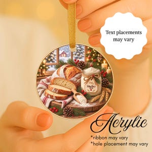May include: A round acrylic Christmas ornament featuring a festive holiday scene with a loaf of bread, sliced bread, a jar labeled Shelley 2026, pine branches, and red berries. The ornament hangs from a gold ribbon against a blurred background.