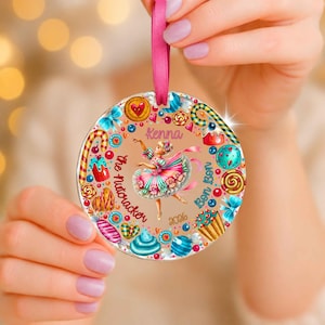 May include: A round, flat, beige-colored ornament featuring a central illustration of a ballerina in a colorful dress. The design includes candy, cupcakes, and lollipops, with the text "Kenna", "the Nutcracker", "Bon Bon", and "2024" printed in script.