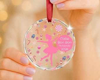 May include: A clear circular acrylic ornament featuring a pink silhouette of a ballerina, a pink circle with the text Callie Bon Bon The Nutcracker 2026, and a pattern of candy canes, cupcakes, lollipops, and nutcrackers, suspended by a red ribbon.