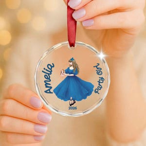May include: A clear circular acrylic ornament featuring a printed illustration of a girl in a blue dress holding a small doll. The text reads Amelia, Party Girl, and 2026 in blue script. A red ribbon is attached to the top for hanging purposes.