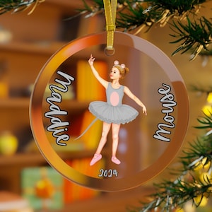 May include: A clear, round Christmas ornament with a gold ribbon. The ornament features a ballerina mouse design with the name "Maddie" and the word "Mouse" in script. The ballerina wears a gray tutu and pink shoes, with the year 2024.