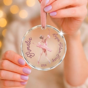 Peut inclure: Une décoration en verre circulaire et transparente ornée d'une illustration de ballerine rose au centre. Le texte Amelia est imprimé à gauche, Sugar Plum Fairy à droite et 2026 en bas en écriture rose foncé. Elle est suspendue par un ruban de velours rose.