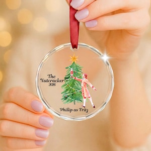 May include: A clear circular acrylic ornament featuring a watercolor illustration of a green Christmas tree and a figure in a red coat. The text reads The Nutcracker 2026 and Phillip as Fritz. A red satin ribbon is attached to the top for hanging.