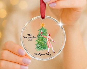 May include: A clear circular acrylic ornament featuring a watercolor illustration of a green Christmas tree and a figure in a red coat. The text reads The Nutcracker 2026 and Phillip as Fritz. A red satin ribbon is attached to the top for hanging.
