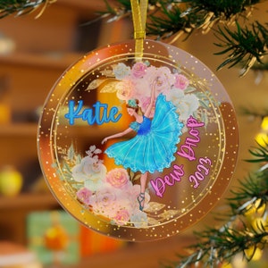 May include: A round, clear acrylic ornament featuring a ballerina in a blue tutu surrounded by pink and white floral illustrations. The text Katie is printed in blue, and the text Dew Drop 2023 is printed in pink. A gold ribbon is attached to the top.