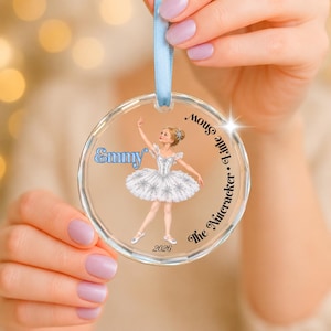 Peut inclure: Une décoration circulaire transparente en acrylique suspendue par un ruban bleu clair. L'ornement présente une illustration imprimée d'une ballerine en tutu blanc, le prénom Emmy, le texte The Nutcracker Little Snow et l'année 2026 en noir.