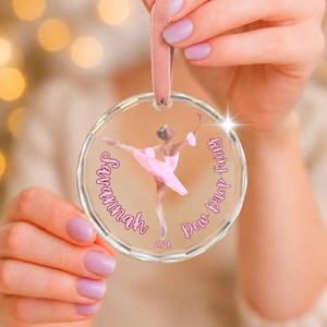 May include: A clear circular acrylic ornament featuring a ballerina in a pink tutu. The text reads Savannah and Dew Drop Fairy with the year 2026 printed below. The ornament is suspended by a soft pink velvet ribbon for hanging on a tree or display.