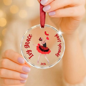 May include: A clear circular acrylic ornament hanging from a red ribbon. The center features a graphic of a dancer in a red and black dress holding a red fan. The text reads Ivy Grace, Spanish Lead, and 2026 in red script lettering.