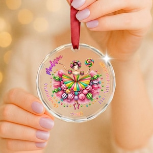 May include: A clear circular acrylic ornament featuring a colorful illustration of a ballerina in a candy-themed tutu. The text reads Madison, The Nutcracker, and 2026. The ornament is suspended by a dark red satin ribbon for hanging on a tree.