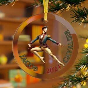 May include: A round glass ornament with a gold rim and a picture of a male dancer in a green and gold costume. The ornament also has the text "Jacob Cavalier 2024" printed on it.