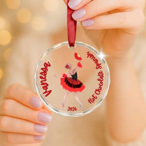 May include: A clear circular acrylic ornament featuring a red ribbon hanger. The design includes a graphic of a dancer in a red and black tutu holding a fan, with the red text Vanessa, Hot Chocolate Spanish, and the year 2026 printed on it.