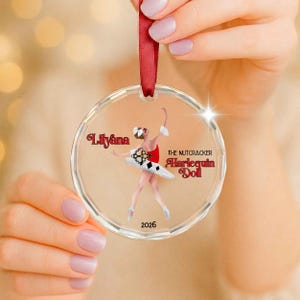 May include: A clear circular acrylic ornament featuring a printed illustration of a ballerina in a red and white harlequin-patterned tutu. The text reads Lilyana, The Nutcracker Harlequin Doll, and 2026. It hangs from a dark red satin ribbon.