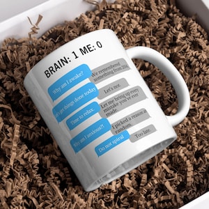 May include: White ceramic mug with a curved handle, featuring text bubbles with phrases like "Why am I awake?" and "Do not spiral." The mug's design includes the text "BRAIN: 1 ME: 0."