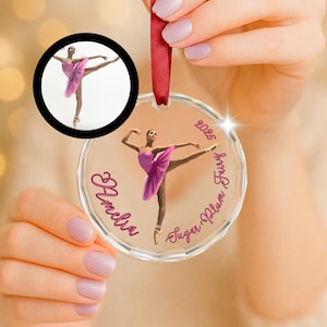 May include: A clear circular acrylic ornament featuring a printed image of a ballerina in a pink tutu, the name Amelia, the text Sugar Plum Fairy, and the year 2026 in pink script. A red ribbon is attached to the top for hanging purposes.