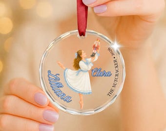 May include: A clear circular acrylic ornament hanging from a red ribbon, featuring an illustration of a girl in a white dress holding a nutcracker. The text reads Lilliana, Clara, and The Nutcracker 2026 in blue and black script lettering.
