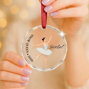 Peut inclure: Une décoration circulaire transparente en acrylique ornée d'une illustration imprimée de ballerine en tutu blanc. Le texte indique SNOW QUEEN, 2026, et le prénom Isabel. Un ruban de satin rouge est fixé au sommet pour suspendre l'objet.