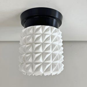 May include: A modern ceiling light fixture with a geometric design. The light has a black base and a white, textured shade with a repeating diamond pattern. The fixture is designed to be mounted on a ceiling.