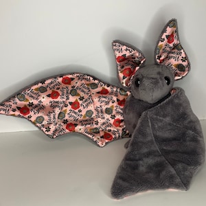 May include: A plush gray bat with large ears and wings. The wings and ear interiors feature a pink floral pattern with the words "Nasty Woman" in black script. The bat has black eyes and a soft, textured body.