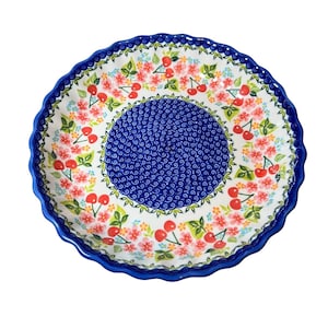 May include: A white ceramic pie plate with a blue and white swirl pattern in the center. The rim of the plate is decorated with a floral pattern featuring pink flowers and red cherries.