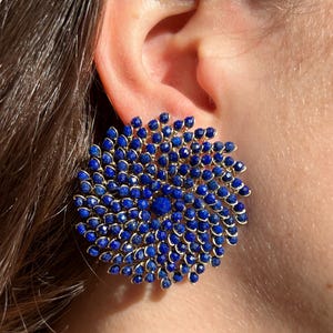 Könnte beinhalten: Nahaufnahme eines großen, runden Ohrrings mit goldfarbenem Rahmen. Der Ohrring ist mit kleinen, tiefblauen Edelsteinen bedeckt, die in einem radialen Muster angeordnet sind. Der Ohrring wird an einem Ohr getragen.